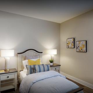 A cozy bedroom featuring a single bed with decorative pillows, flanked by two nightstands with lamps. The room is adorned with wall art for an added touch of style.