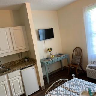 A cozy room with a kitchenette, featuring a bed, wall-mounted TV, desk, chair, and a window.