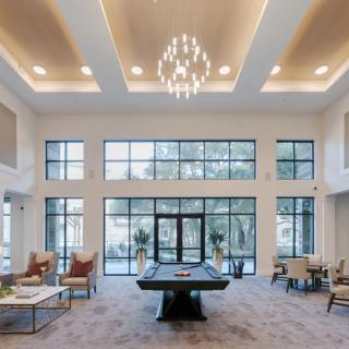 A modern lobby with a billiards table, seating areas, and a large chandelier, illuminated by natural light from expansive windows.