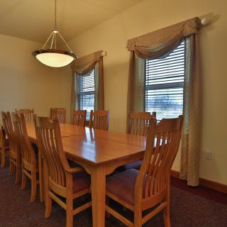 A spacious dining room with a large wooden table and several chairs, lit by a chandelier and natural light from the windows.