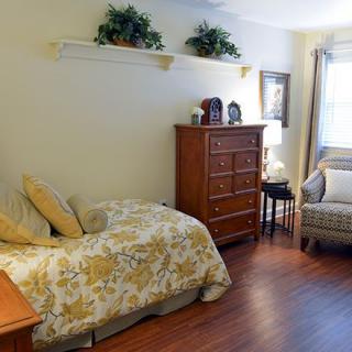A cozy bedroom with a wooden floor, featuring a bed, dresser, and armchair, beautifully decorated with plants and art.