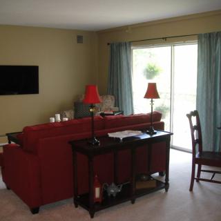 A cozy living room with a red sofa, lamps, and a television mounted on the wall. The room has a sliding glass door with blue curtains.