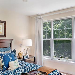 A cozy bedroom with a large window, decorated in blue and white tones, featuring a bed with patterned bedding and a bedside lamp.