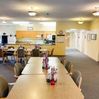 A spacious and well-lit dining area with multiple tables and chairs, decorated with small centerpieces, adjacent to a kitchen space.