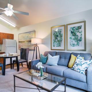 A stylish and cozy living room featuring a comfortable sofa with colorful cushions, framed artwork on the wall, and a modern coffee table. A dining area is visible in the background.