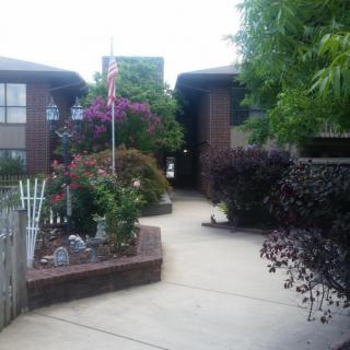 A well-maintained garden pathway leading to a brick building, adorned with vibrant plants and a flag.