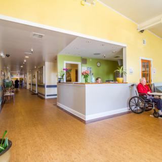 A bright and welcoming nursing home reception area with a person in a wheelchair and a view down the hallway.
