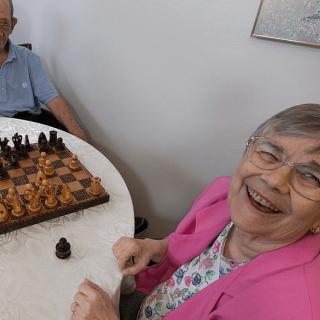 An elderly woman smiles while playing a game of chess at a table.