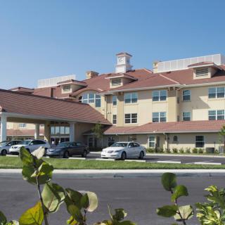 A mid-sized hotel with a brown roof, featuring parked cars and surrounding greenery under a clear blue sky.