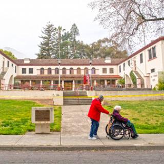 A caregiver pushes an elderly person in a wheelchair outside a residential building.