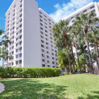A high-rise apartment building with palm trees and a grassy area in the foreground under a clear blue sky.