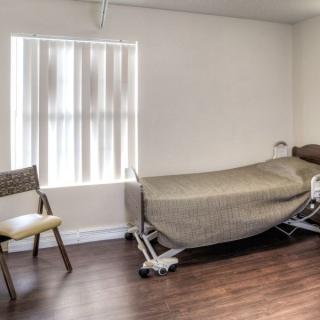 A minimalist room with a hospital bed and chair, featuring a wooden floor and window with vertical blinds.