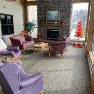 A cozy living room with purple armchairs facing a stone fireplace, decorated with a red Christmas tree near large windows.