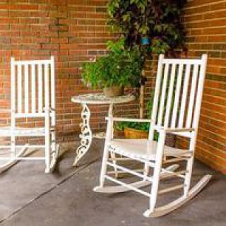 Two white rocking chairs and a small table adorn a cozy patio area against a brick wall, accented by lush greenery.