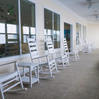 A row of white rocking chairs lined up along a porch with large windows.