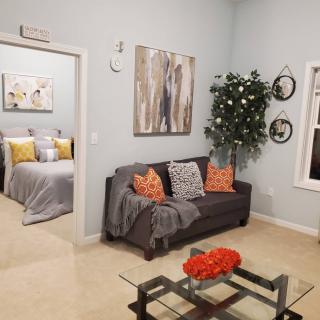 A cozy living room with a gray sofa, colorful pillows, and modern artwork. A glimpse into the bedroom is visible through an open doorway.