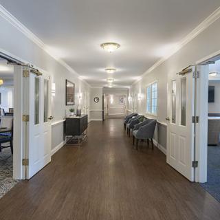 A well-lit hallway with wooden flooring and multiple doors, featuring chairs arranged along the side.