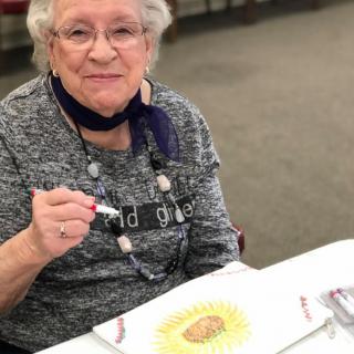 A smiling elderly woman engaged in an art activity, holding a marker and working on a colorful drawing.