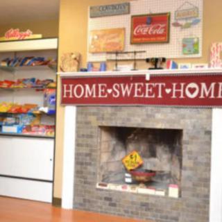A cozy interior featuring a 'Home Sweet Home' sign above a fireplace, with shelves of food items and vintage Coca-Cola signage.