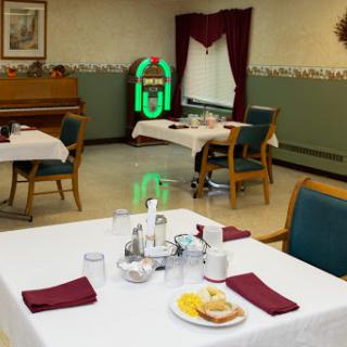 A cozy dining area set for a meal, featuring tables with white tablecloths and food items, alongside a piano in the background.