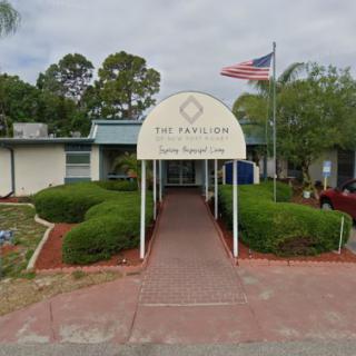 An entrance to a single-story building with a pathway, surrounded by greenery and an American flag nearby.
