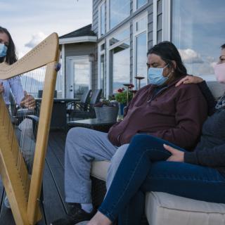 A group of people wearing masks sit outside while one person plays a harp.