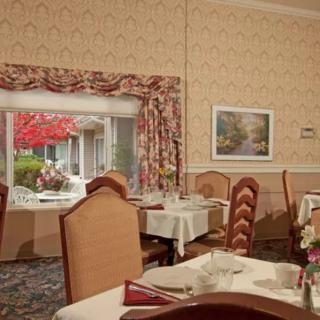A cozy dining area with neatly set tables and upholstered chairs, featuring a large window with floral curtains and decorative artwork on the walls.