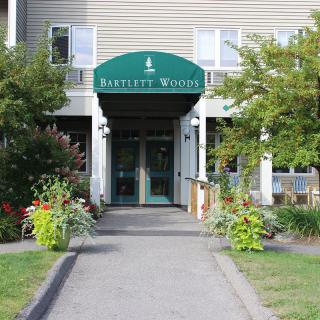 The entrance of a building with a green awning labeled 'Bartlett Woods', surrounded by plants and trees.