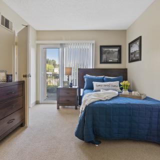 A cozy bedroom with a neatly made bed and blue bedding, featuring a lamp on a bedside table and natural light streaming through the window.
