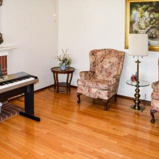 A cozy living room with a digital piano, two floral armchairs, a lamp, and a painting on the wall.
