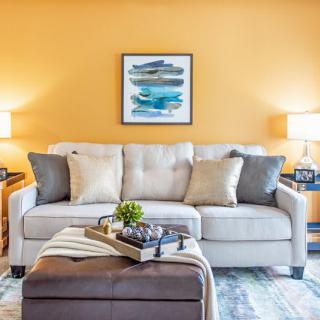 A cozy living room with a light gray sofa, accented by colorful cushions, set against a vibrant yellow wall featuring modern artwork. Two side lamps provide warm lighting, complementing the stylish decor.