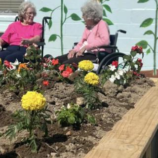 Two elderly women enjoy a sunny day in the garden, surrounded by colorful flowers.