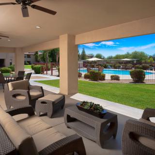 A covered patio with comfortable seating overlooks a pool and landscaped garden.