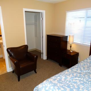 A tidy bedroom featuring a leather chair, a dresser with a lamp, an open closet, and a window.