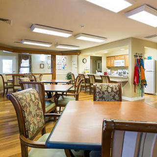 A well-lit dining room with multiple tables and chairs, adjacent to a kitchen area in a community space.