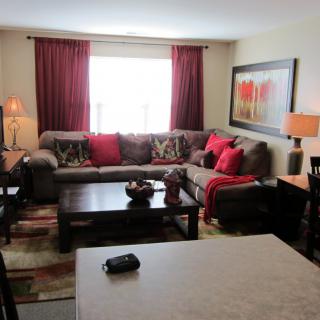 A cozy living room with a large sectional sofa, accented by red pillows and curtains, featuring a coffee table and decorative lamps.