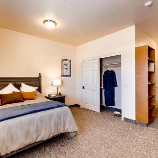 A cozy bedroom featuring a large bed, a lamp on a bedside table, a partially open closet, and carpeted flooring.