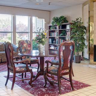 A cozy seating area with classic wooden furniture and a bookshelf, surrounded by potted plants and a large window.
