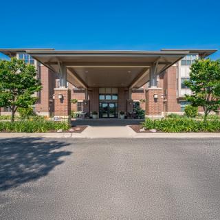 The image shows the front entrance of a modern building with a covered driveway, surrounded by well-maintained landscaping and trees under a clear blue sky.