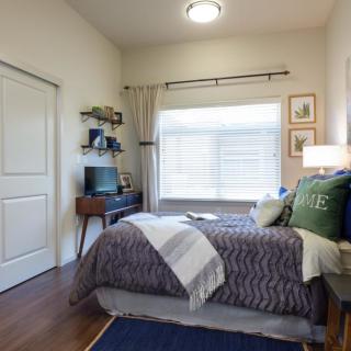 A cozy bedroom with modern decor, featuring a neatly made bed, a small desk with shelves, and a large window with curtains.