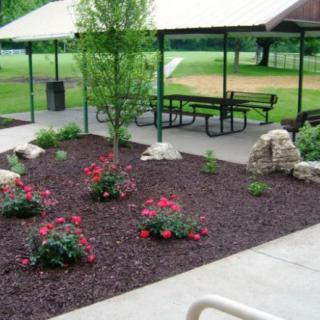 A landscaped park area featuring a pavilion with picnic tables, surrounded by flower beds and mulch.