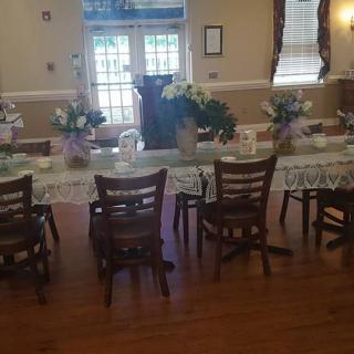 A decorated dining room with a long table set for an event, featuring floral arrangements and wooden chairs.