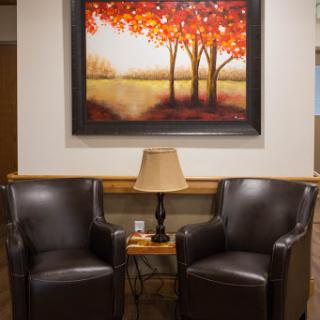 A cozy seating area featuring two leather chairs, a side table with a lamp, and a vibrant painting of trees in autumn colors.
