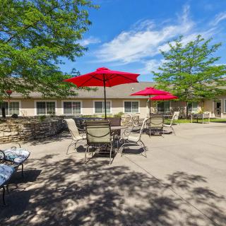 A sunny outdoor patio area with tables, chairs, and red umbrellas near a building.