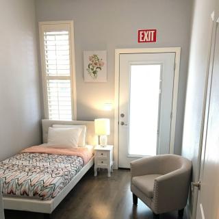 A minimalist bedroom with a single bed, a small side table with a lamp, and an armchair. The room is brightened by natural light from the window.