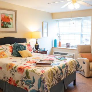 A cozy bedroom with colorful floral bedding, an armchair by the window, and warm lighting.