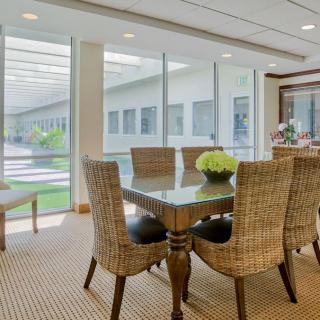 A modern dining room with wicker chairs and a glass table, well-lit by large windows overlooking a garden.