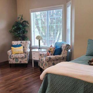 A cozy bedroom with two patterned chairs, a small round table between them, a bed with teal bedding, and a large window allowing natural light.