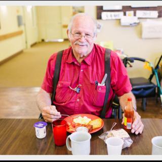 An elderly man in a red shirt enjoying a meal at a dining table indoors.