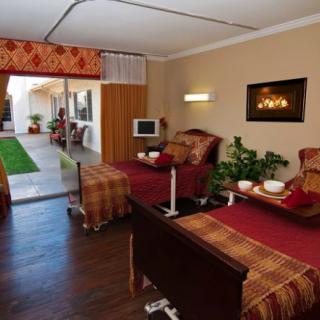 A cozy room featuring two beds, a mounted television, and an electric fireplace, with large windows overlooking a garden.